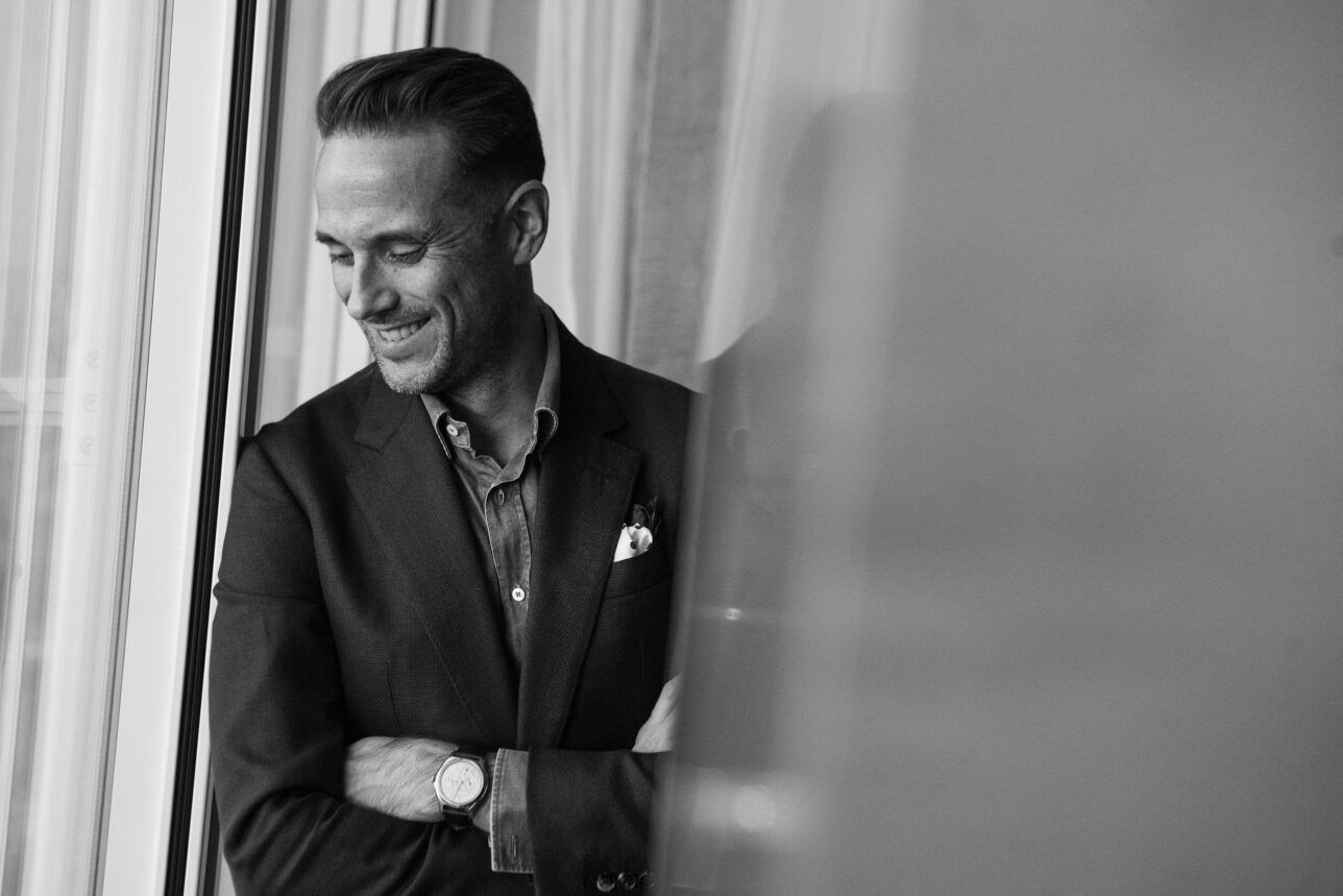 Dark textured sport coat with chambray shirt and dotted pocket square, worn by a model by a window in black and white
