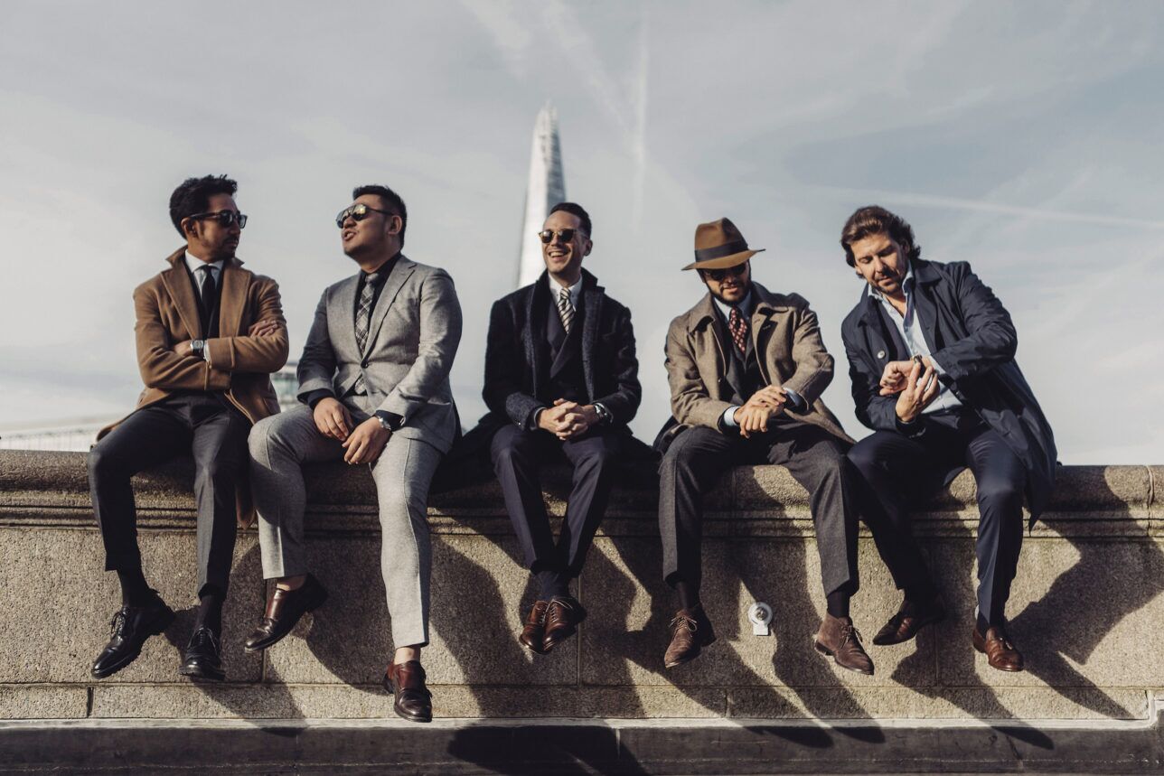 Tailored overcoats, tweed and flannel suits with neckties, worn by five men on a stone wall