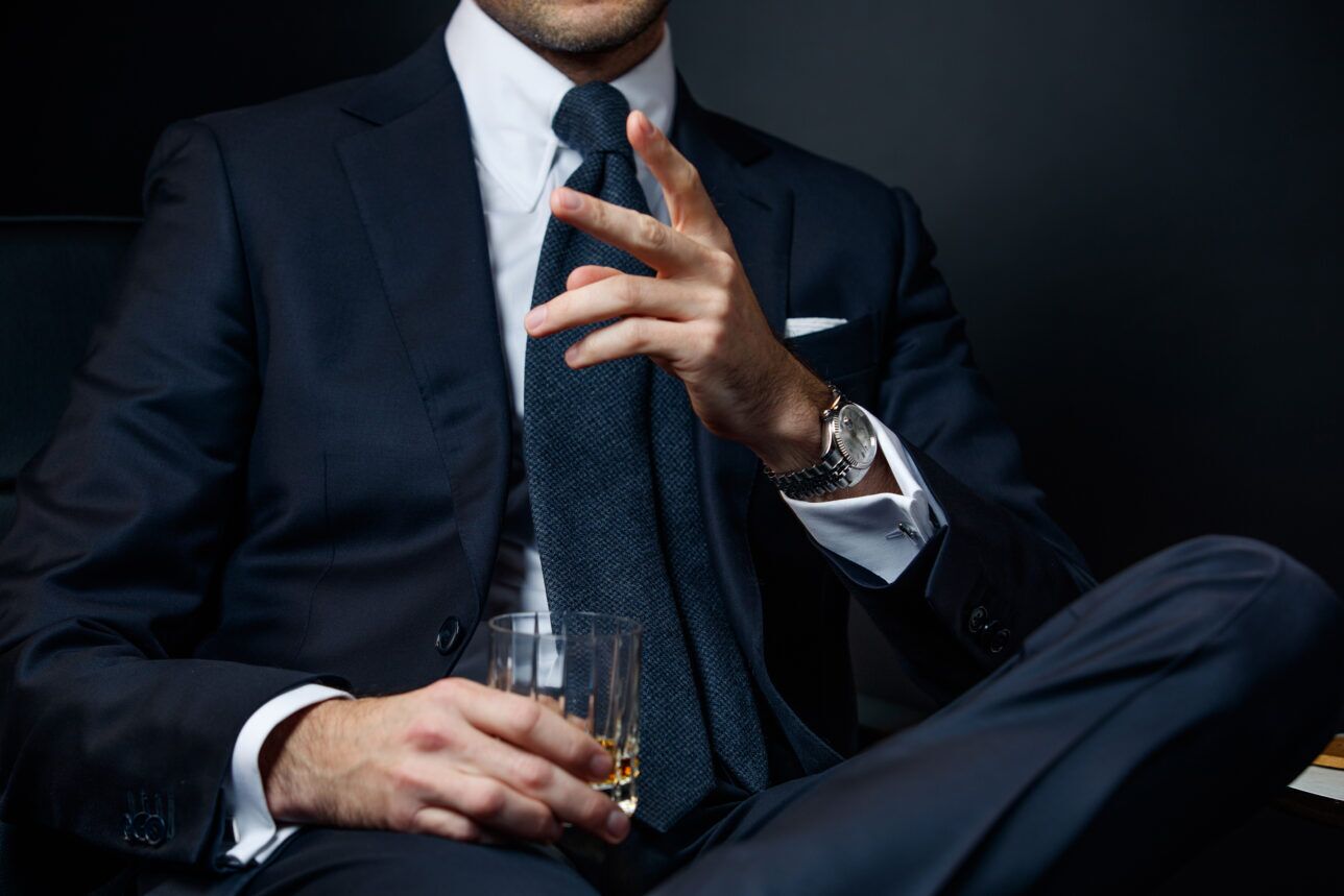Navy suit with white shirt, blue textured tie and white pocket square, worn by a seated model with whisky