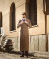 Brown glen check double-breasted wool overcoat with white shirt and black tie, worn by a model on a cobbled street