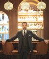Charcoal pinstripe three-piece suit with white shirt and striped tie, worn by a model in a hotel bar