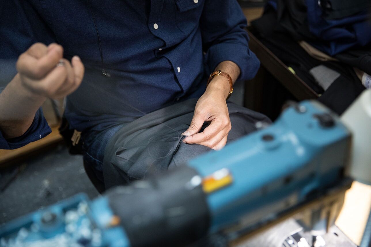 Grey wool trouser fabric being hand-stitched by a tailor at a sewing machine, a workshop detail