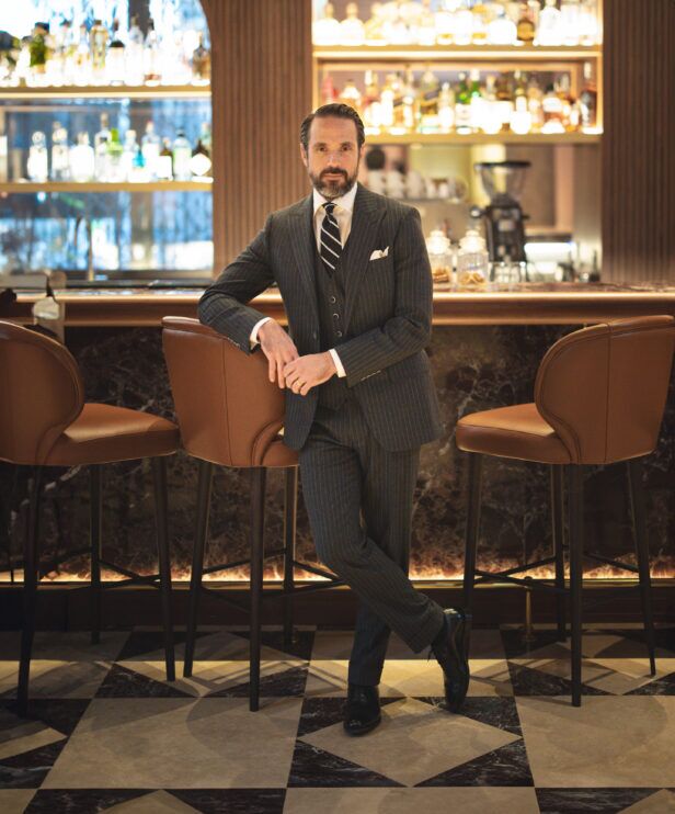 Charcoal pinstripe three-piece wool suit with white shirt, striped tie and white pocket square, worn by a model at a bar