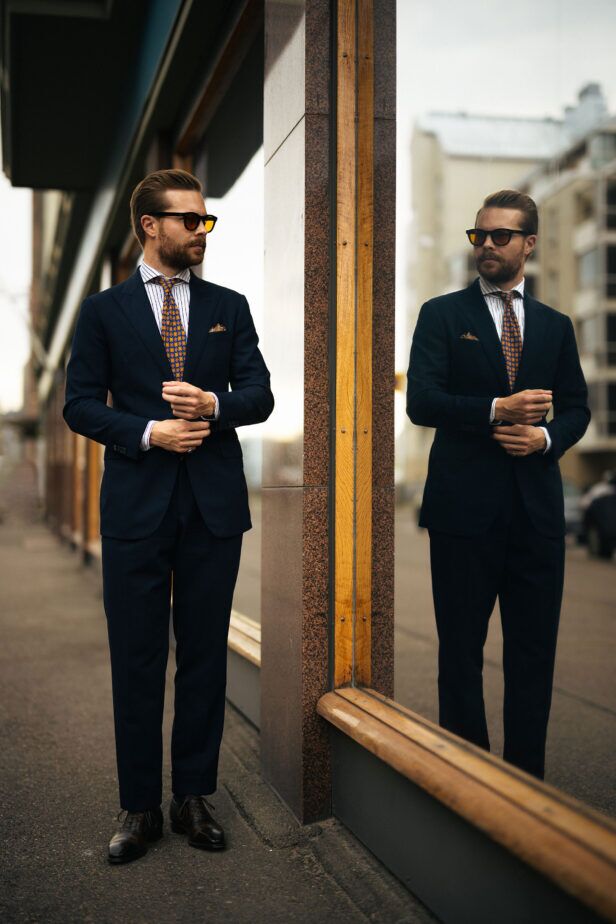Navy two-piece suit with striped shirt, orange medallion silk tie and brown oxfords, worn by a model in a styled street scene