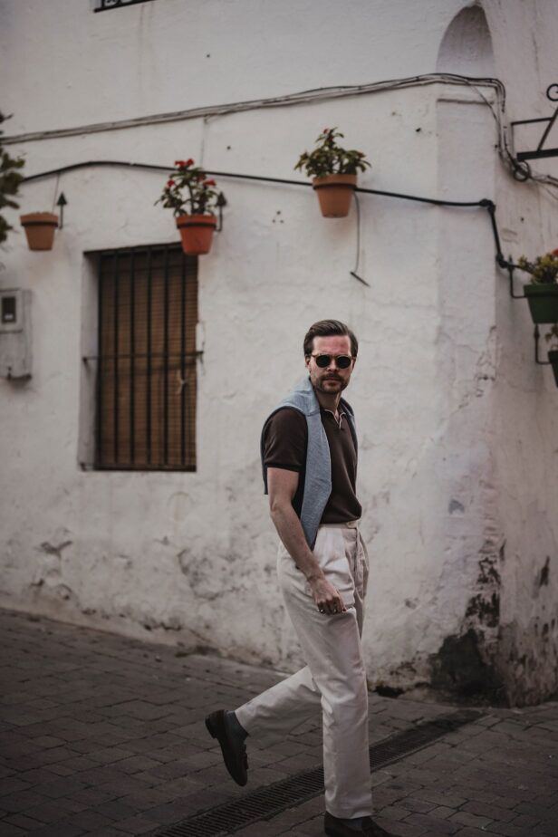 Brown knit short-sleeve polo with cream pleated trousers and brown loafers, worn by a model on a whitewashed street