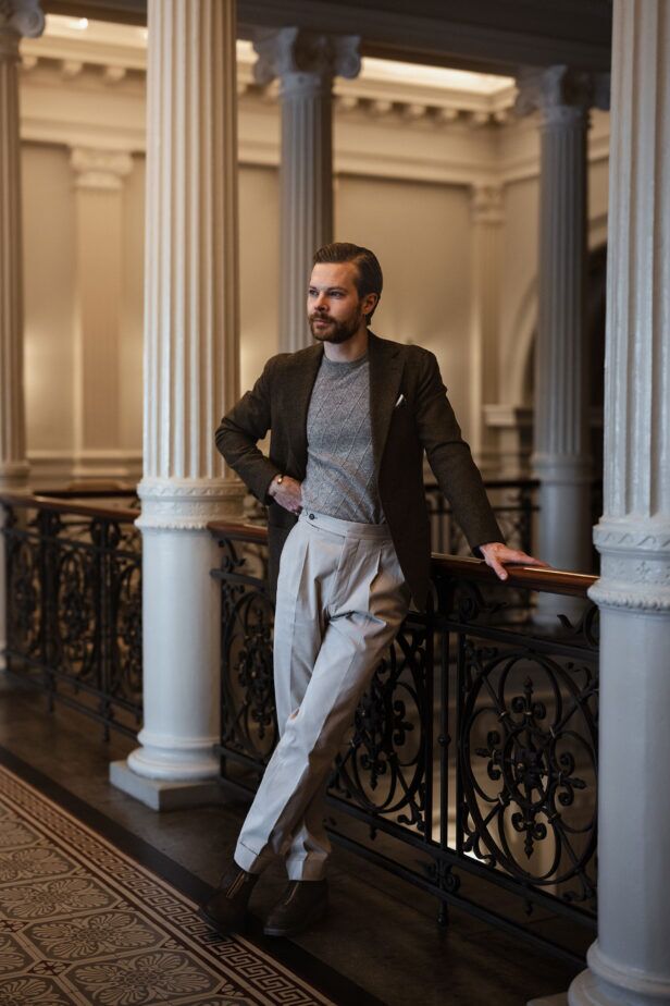 Olive brown wool sport coat with grey diamond-knit crewneck and cream pleated trousers, worn indoors