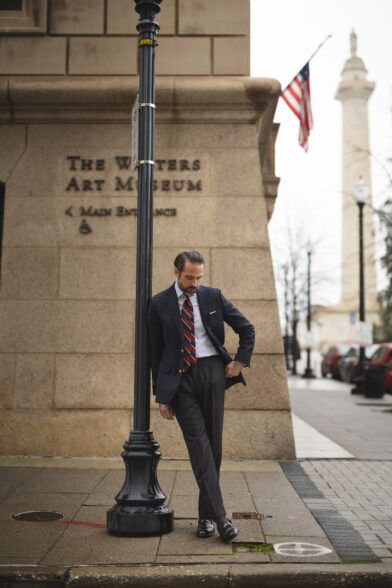 Navy blazer with gold buttons, burgundy striped tie, white shirt and grey trousers, worn by a model on a city street