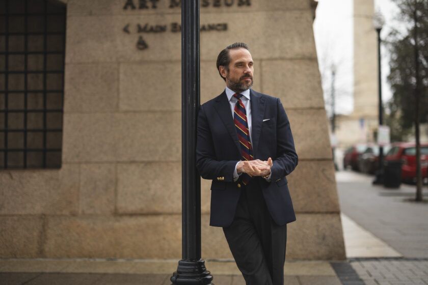 Navy blazer with gold buttons, burgundy striped tie and grey trousers, worn by a model on a city street