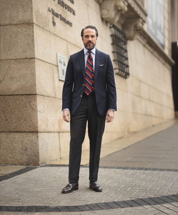 Navy blazer with gold buttons, burgundy striped tie and charcoal flannel trousers, worn by a model on a city street