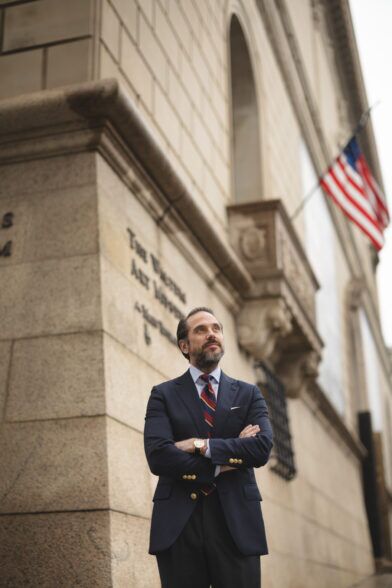 Navy blazer with gold buttons, blue shirt and burgundy striped tie, worn by a model outside a stone museum