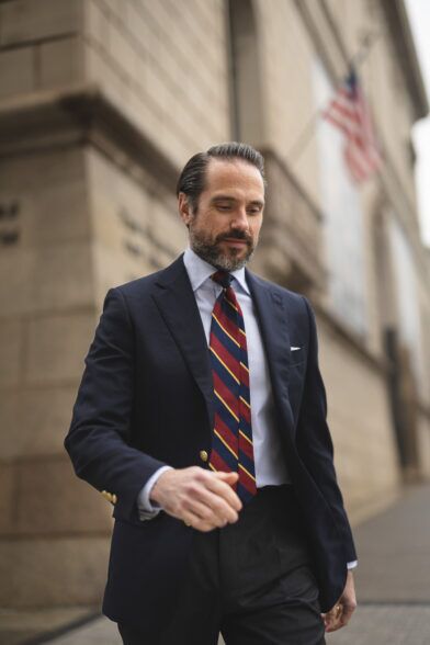 Navy blazer with gold buttons, burgundy and navy repp stripe tie and white shirt, worn by a model on a city street