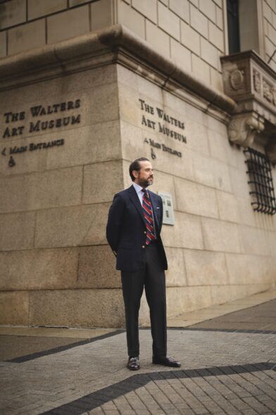 Navy gold-button blazer with blue shirt, red and blue striped tie and charcoal trousers, worn by a model outdoors