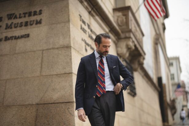 Navy blazer with gold buttons, burgundy and blue striped tie and grey trousers, worn by a model on a city street