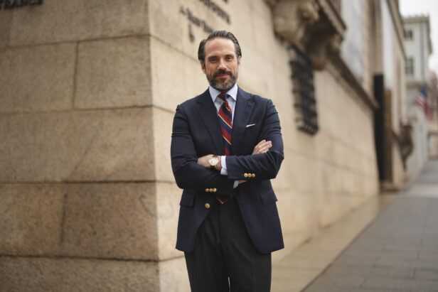 Navy blazer with gold buttons, striped repp tie, blue shirt and grey trousers, worn by a model on a city street