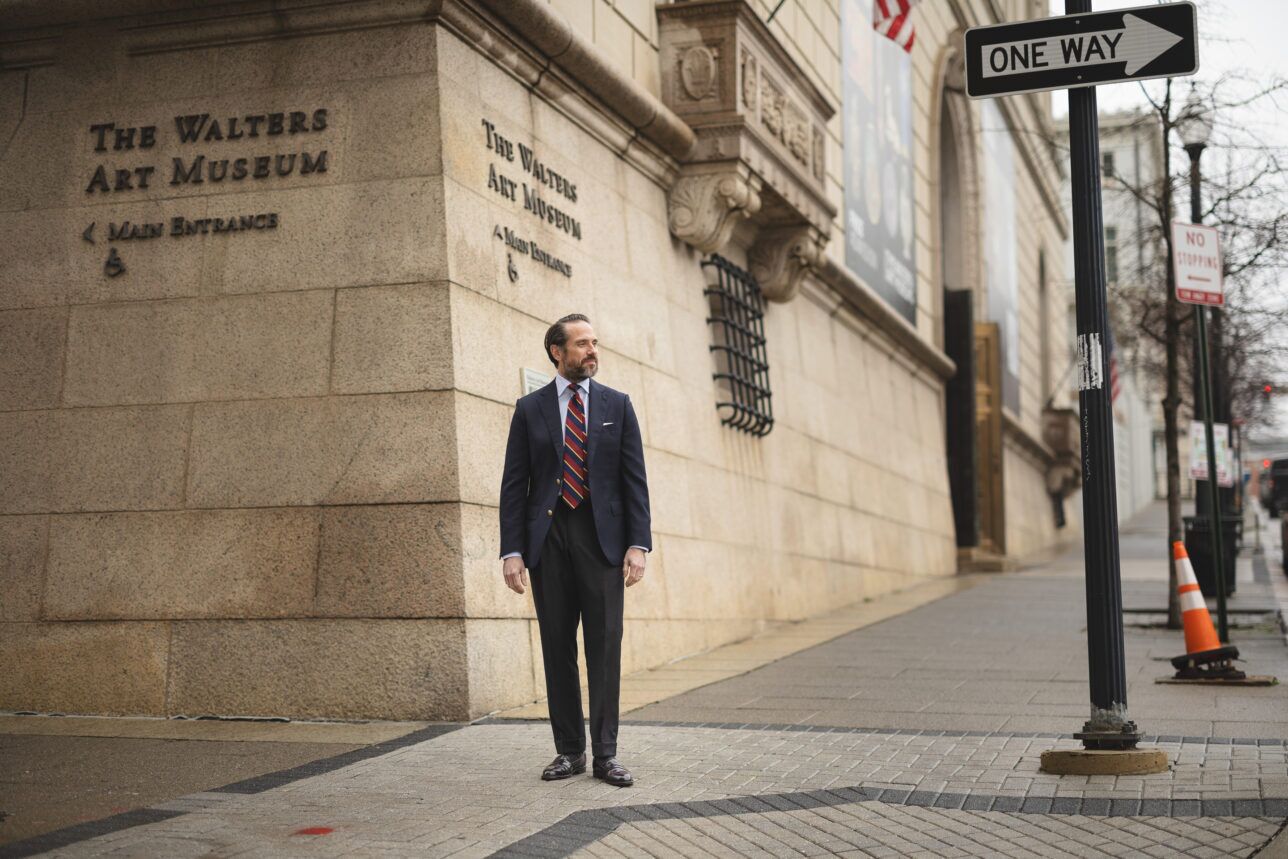 Navy blazer with gold buttons, striped blue shirt, red repp stripe tie and grey trousers, worn by a model on a city street