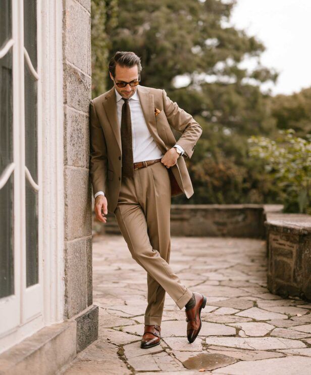 Tan two-button suit with white shirt, brown knit tie and brown double monk shoes, worn by a model outdoors