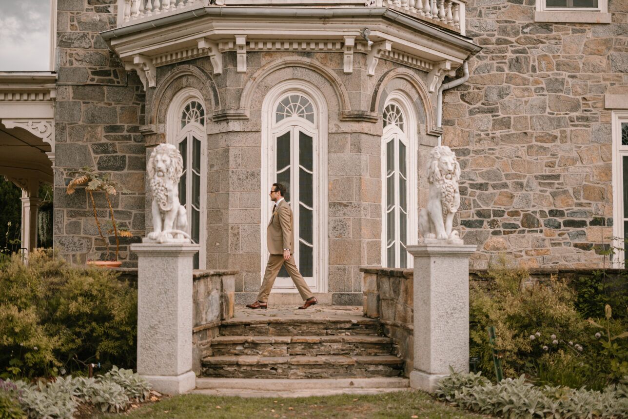 Tan solaro suit with white shirt and brown leather shoes, worn by a model walking past a stone mansion
