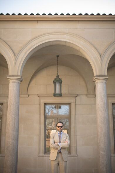 Khaki cotton suit with light blue shirt and striped tie, worn by a model in a stone archway