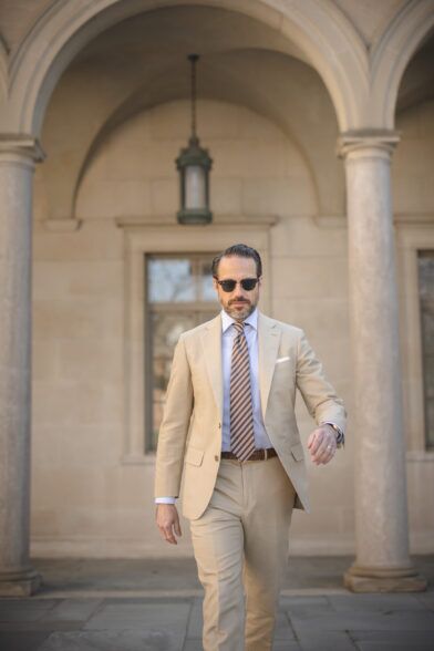 Khaki cotton suit with light blue shirt, brown striped tie and tan belt, worn by a model in a stone arcade