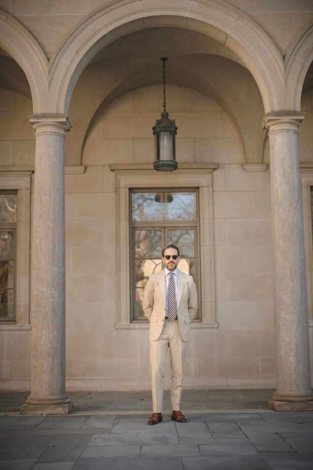Khaki cotton suit with blue striped shirt, brown striped tie and tan monk strap shoes, worn by a model under stone arches