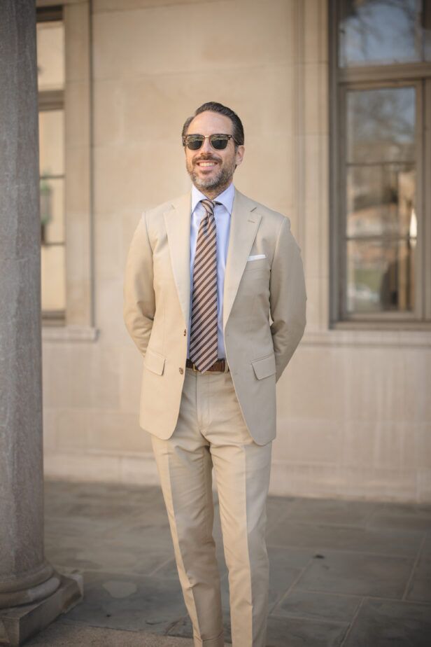 Khaki cotton suit with light blue shirt, brown striped tie and white pocket square, worn by a model outdoors