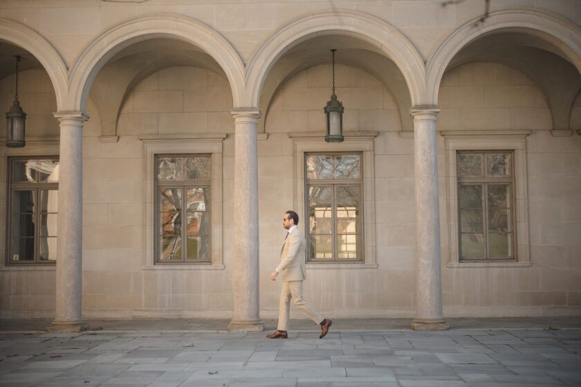 Khaki cotton suit with white shirt and brown leather shoes, worn by a model walking past a stone arcade