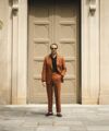 Terracotta linen suit with black open-collar shirt and black loafers, worn by a model outside a stone doorway