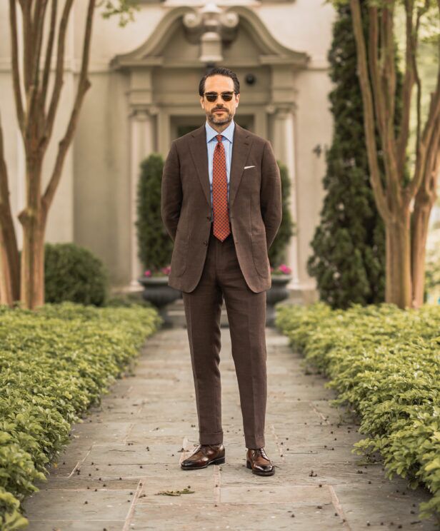 Brown hopsack suit with blue striped shirt, orange medallion tie and brown derbies, worn by a model in a garden