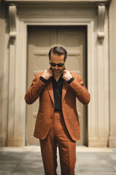Terracotta linen-textured suit with black open-collar shirt, worn by a model on a stone doorstep