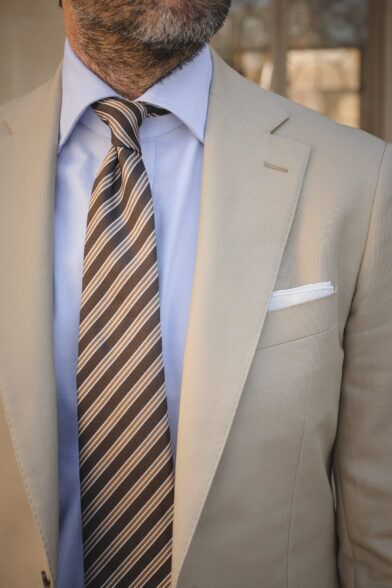 Khaki cotton suit jacket with notch lapel, blue shirt, brown striped silk tie and white pocket square, close-up on a model