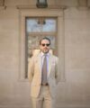 Khaki cotton suit with light blue shirt and brown striped tie, worn by a model outside a stone building