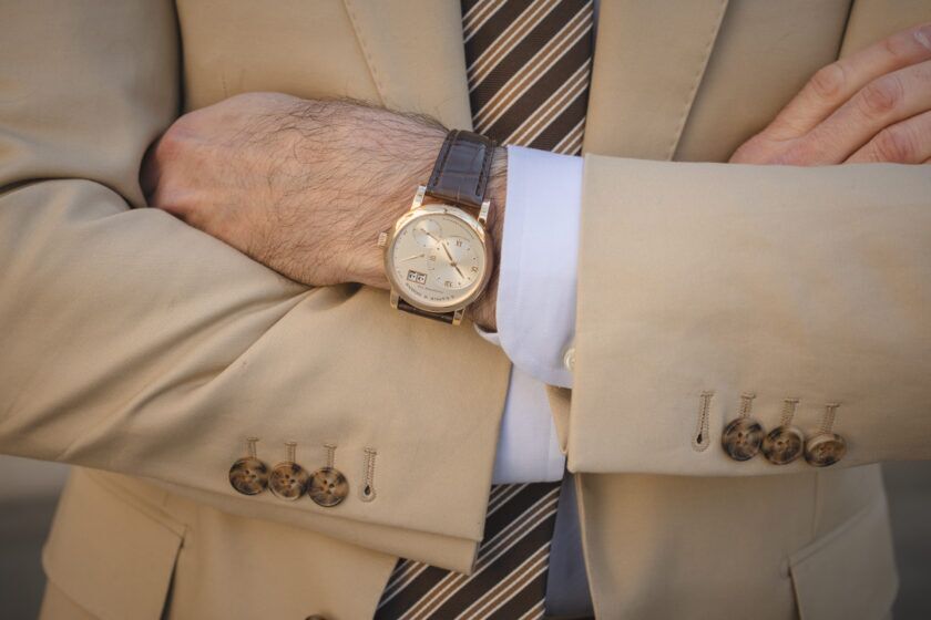 Khaki cotton suit jacket sleeve with horn buttons, white shirt cuff and gold watch on brown strap, a close-up detail