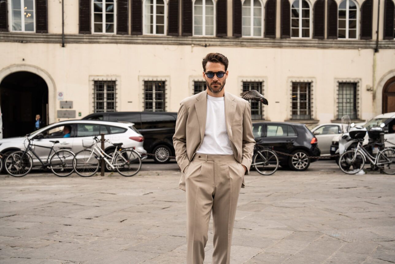 Tan single-breasted suit with pleated trousers and white tee, worn by a model in a European piazza