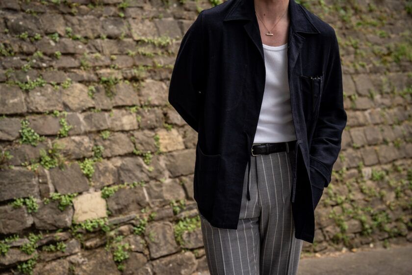Navy patch-pocket chore jacket with white ribbed tank and grey striped trousers, worn by a model against a stone wall