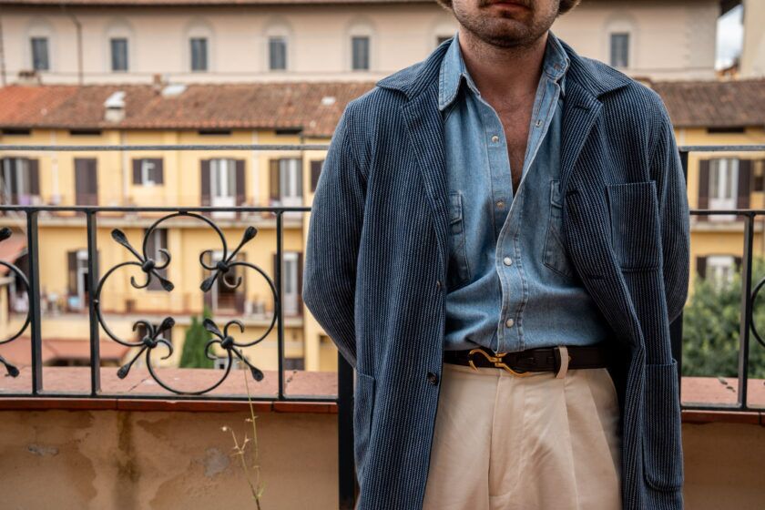 Blue textured corduroy sport coat with denim shirt, brown belt and cream trousers, worn by a model on a balcony