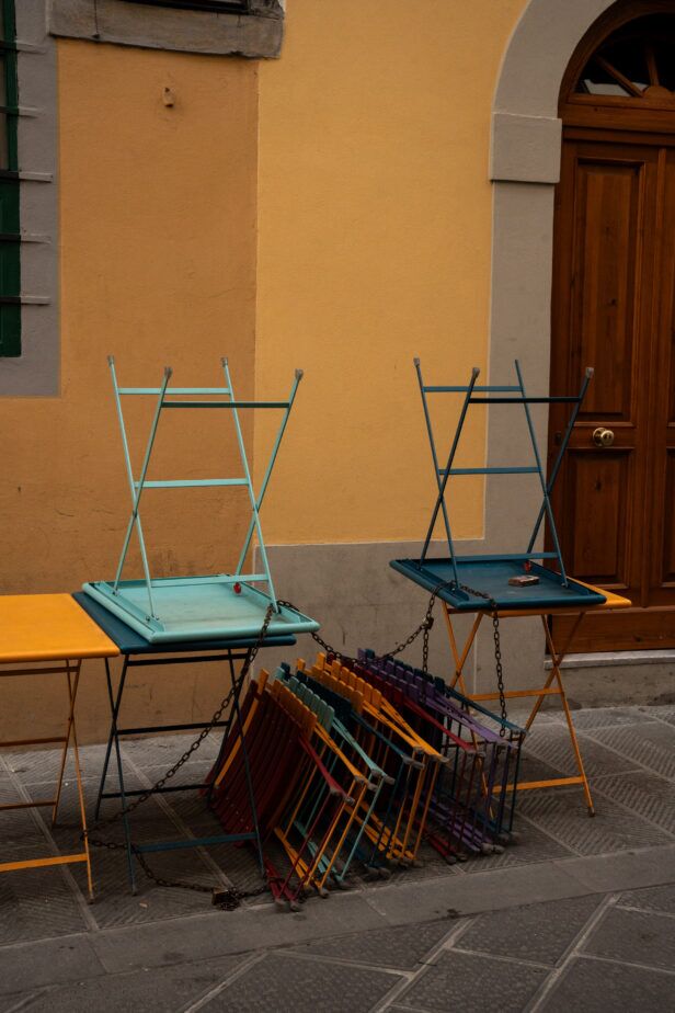 Folded café chairs and tables in teal, red and yellow chained outside a yellow building, a street scene