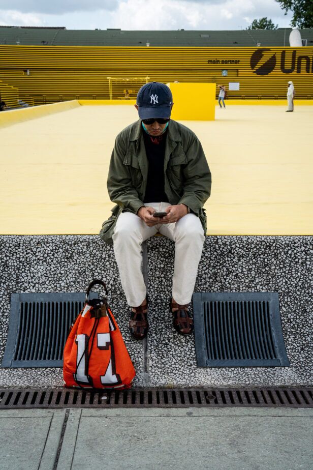 Olive field jacket with cream trousers, navy cap and brown fisherman sandals, worn by a man seated in a styled street scene