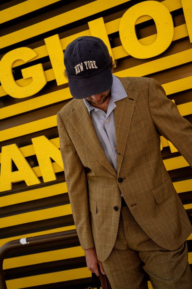 Tan micro-check suit with pale blue shirt and navy New York cap, worn by a model in a styled street scene