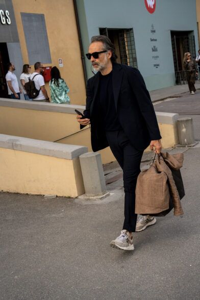 Navy soft-shouldered suit worn with a navy tee and grey sneakers, carrying a tan linen jacket, street scene