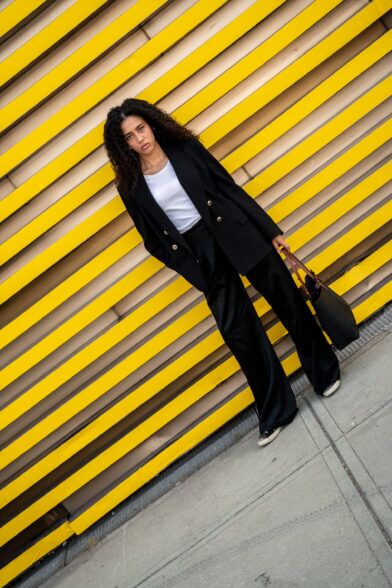 Black double-breasted blazer with white tee and wide-leg black trousers, worn by a model against a yellow wall
