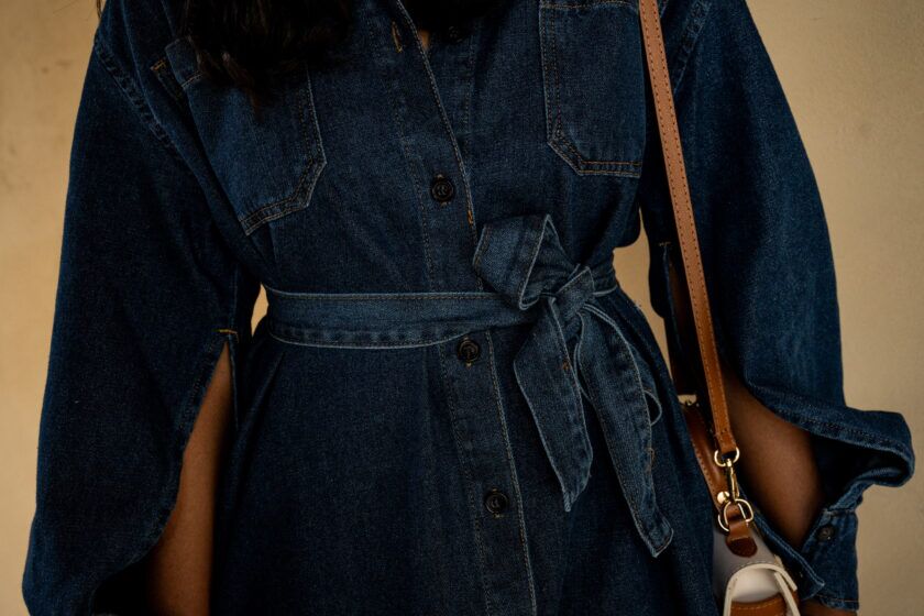 Indigo denim shirt dress with tie belt and chest pockets, worn with a tan leather crossbody strap, detail view