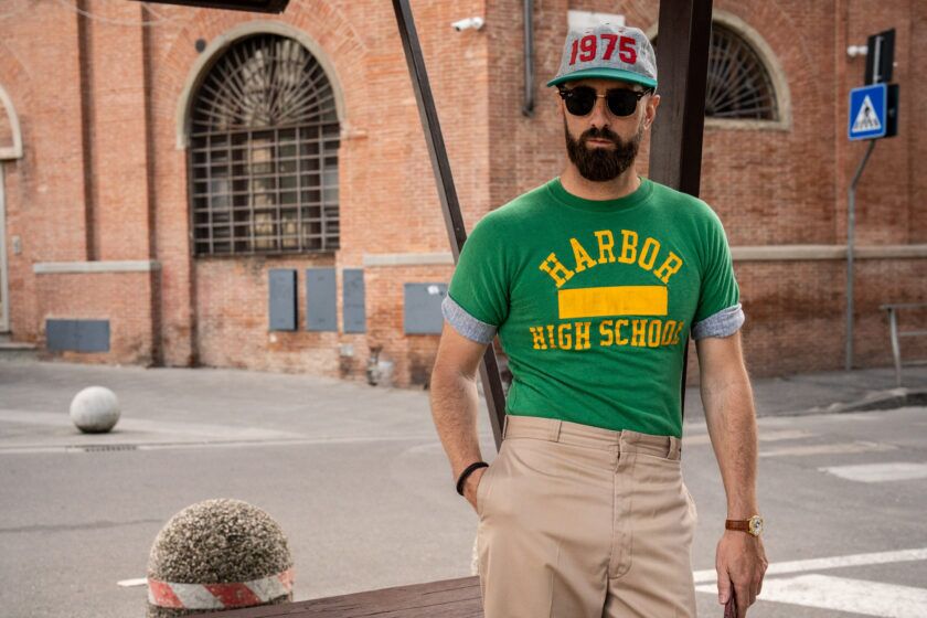 Green graphic T-shirt with yellow varsity print, khaki trousers and a 1975 ball cap, worn on a street