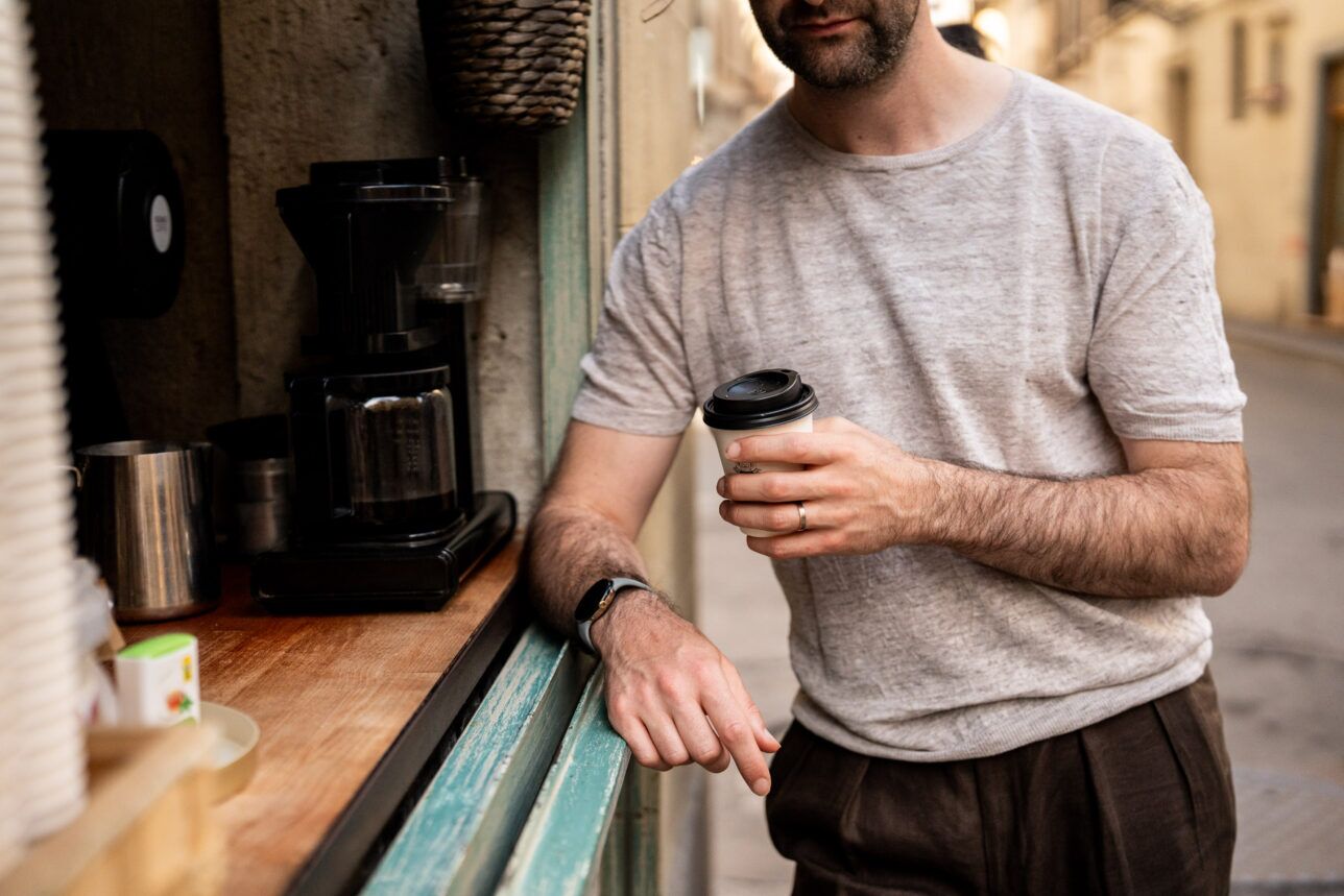 Light grey linen crewneck tee with dark brown pleated trousers, worn by a man at a coffee window in a styled street scene