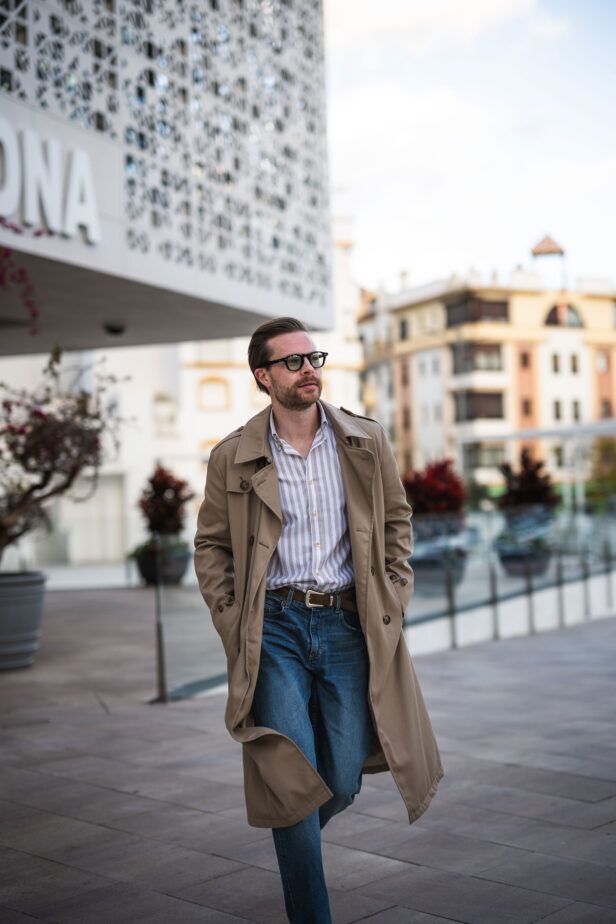 Tan double-breasted trench coat with striped shirt, brown belt and blue jeans, worn by a model on a city street