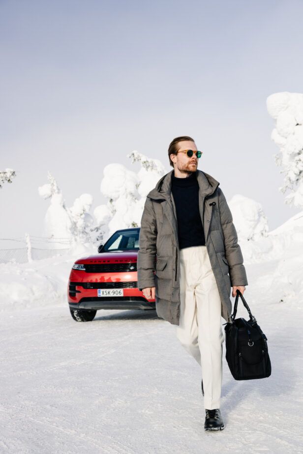 Grey wool down parka with navy roll neck, cream trousers and black holdall, worn by a model in a snowy scene