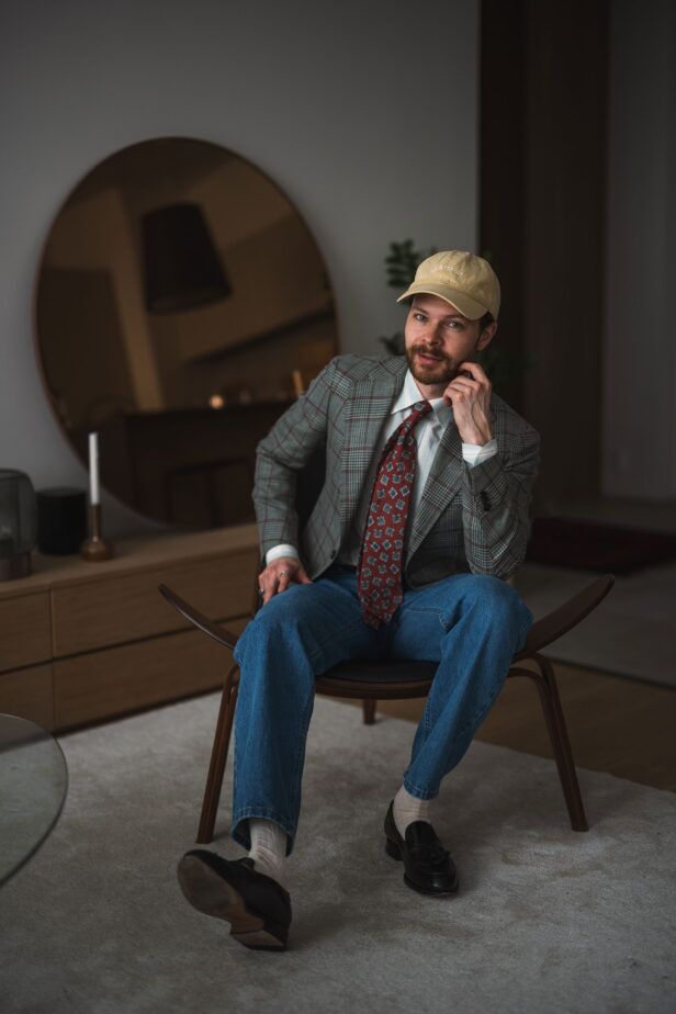 Green glen check sport coat with rust medallion silk tie, blue jeans and black tassel loafers, worn by a seated model