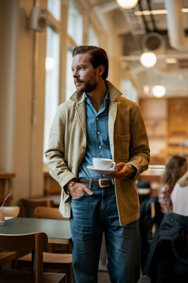 Tan herringbone wool chore jacket with denim shirt, blue jeans and brown belt, worn by a model in a cafe