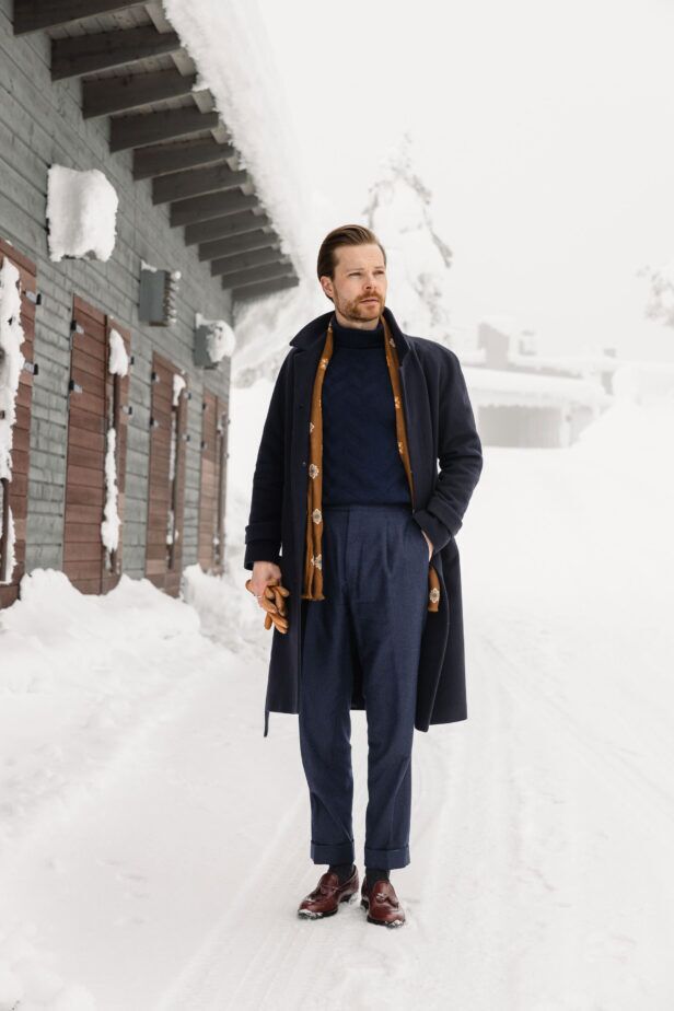 Navy wool overcoat with cable-knit turtleneck, printed scarf and tassel loafers, worn by a model in the snow