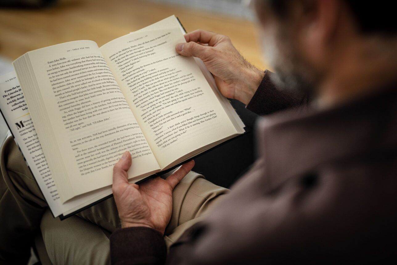 Open hardcover book held by a man in a brown jacket and tan trousers, a styled lifestyle scene