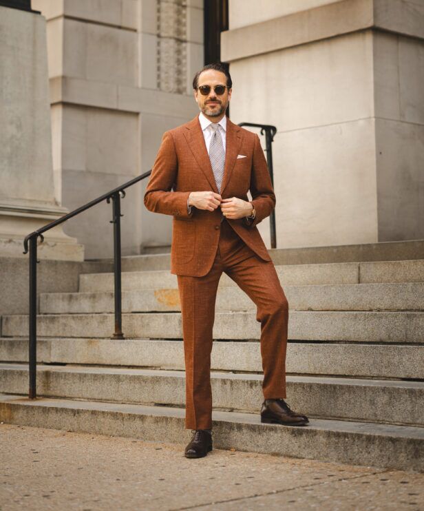 Terracotta linen-blend suit with peak lapels, white shirt, checked tie and brown oxfords, worn by a model on stone steps